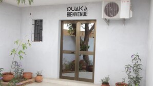 A white building with a sign reading "OUAKA BIENVENUE" above a glass door. There are potted plants around the entrance. - Ouaka Hotel (Ouagadougou)