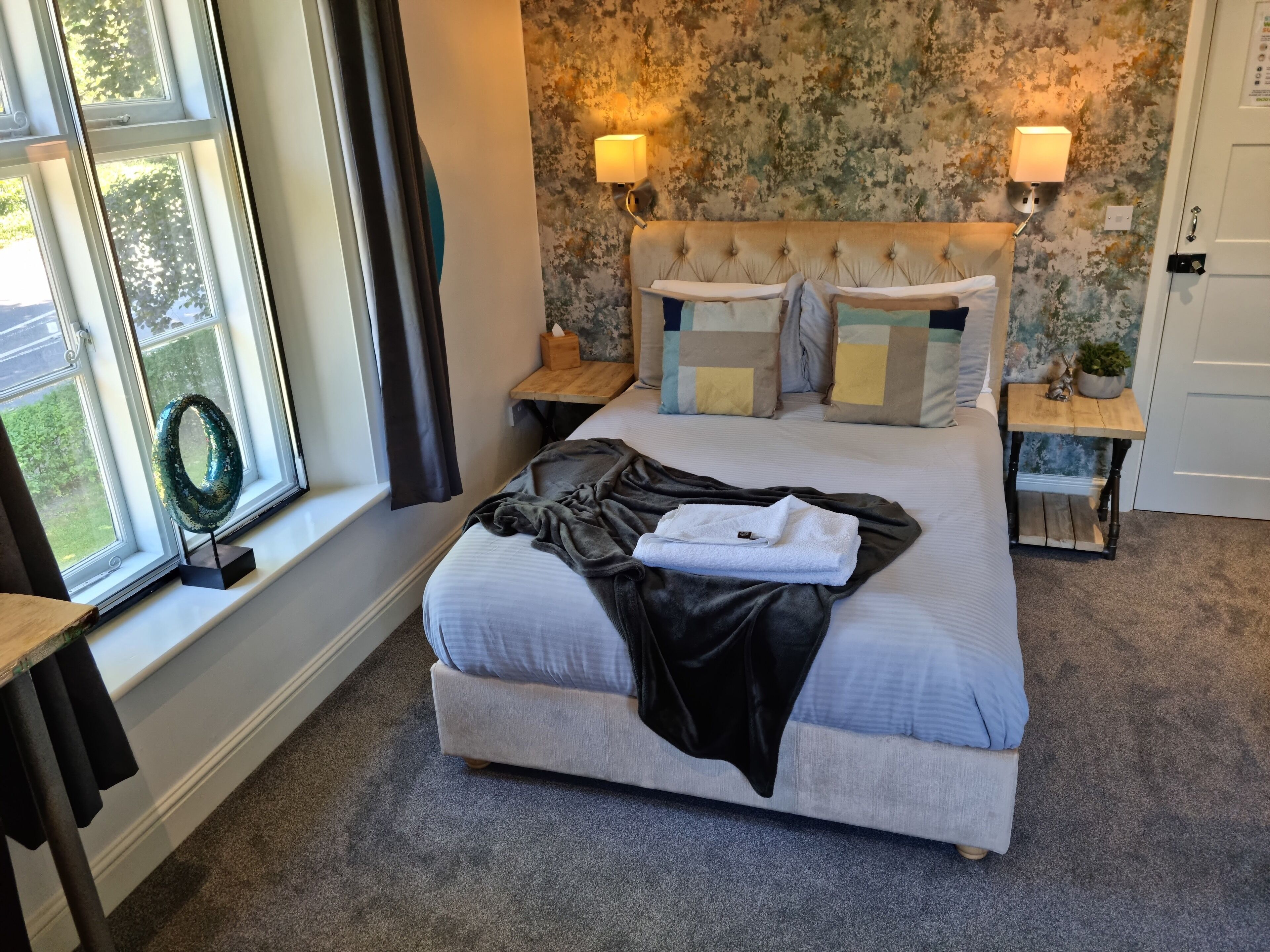 A neatly made bed with a grey blanket and a folded towel, two bedside tables with lamps, a floral wallpaper, and a window with curtains.