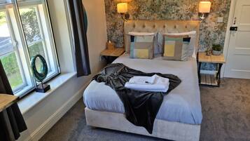 A neatly made bed with a grey blanket and a folded towel, two bedside tables with lamps, a floral wallpaper, and a window with curtains.