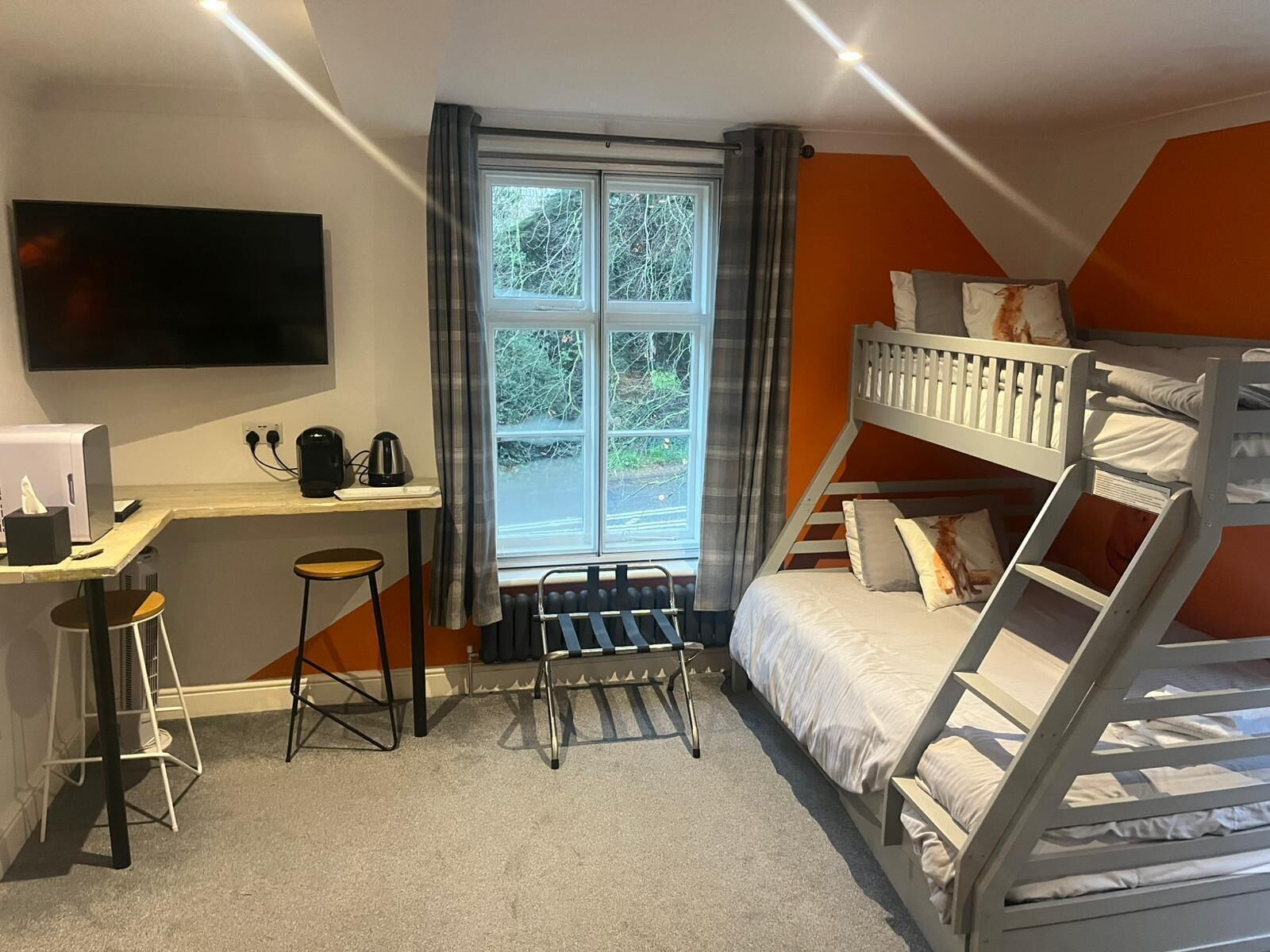 A compact hotel room with a bunk bed, a desk with a computer and a kettle, a TV mounted on the wall, and a window with curtains.
