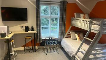 A compact hotel room with a bunk bed, a desk with a computer and a kettle, a TV mounted on the wall, and a window with curtains.