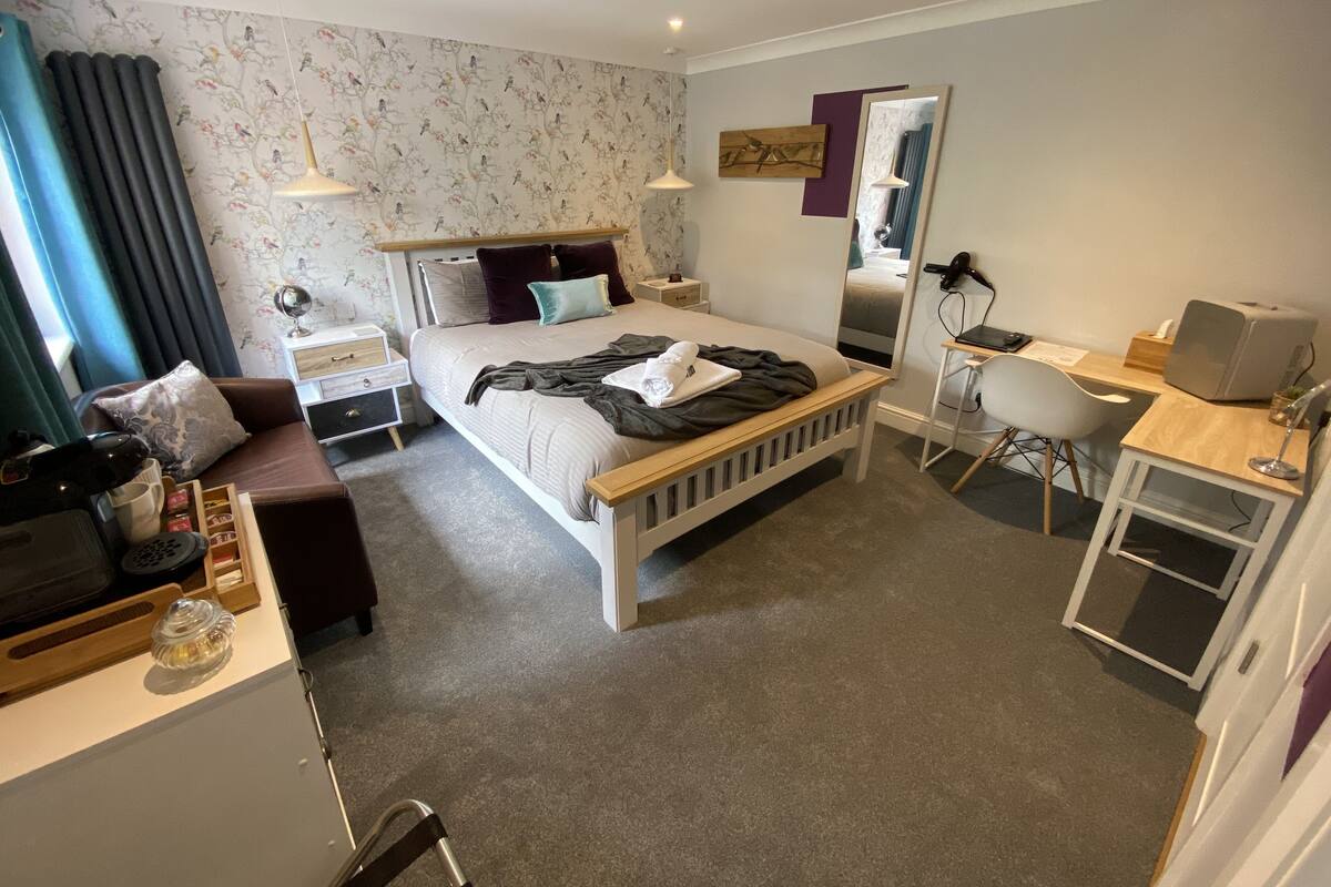 Ett modernt hotellrum med en snyggt bäddad säng, ett skrivbord med en dator, en stol, ett litet bord med en lampa och en spegel.