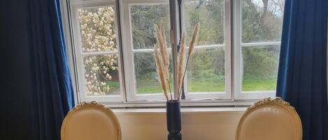 A room with two chairs, a small table with a vase, and a large window with curtains.