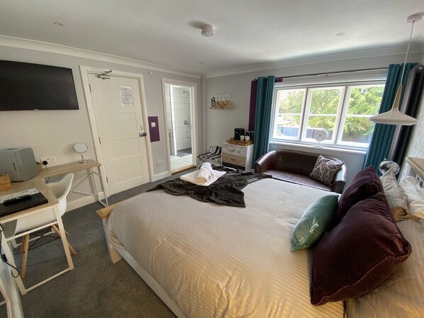 A hotel room with a bed, a desk with a computer, a TV mounted on the wall, a window with curtains, and a small table with a coffee maker. - Copperbeech Bed and Breakfast (Saxmundham)