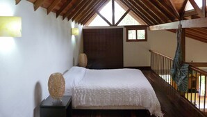 A bedroom with a wooden ceiling, a bed with a white bedspread, a nightstand, a woven basket, and a window with a view of greenery. - Casa Jade (Rionegro)