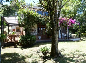 A covered outdoor seating area with a table and chairs, surrounded by lush greenery and a tree with pink flowers. - Casa Jade (Rionegro)