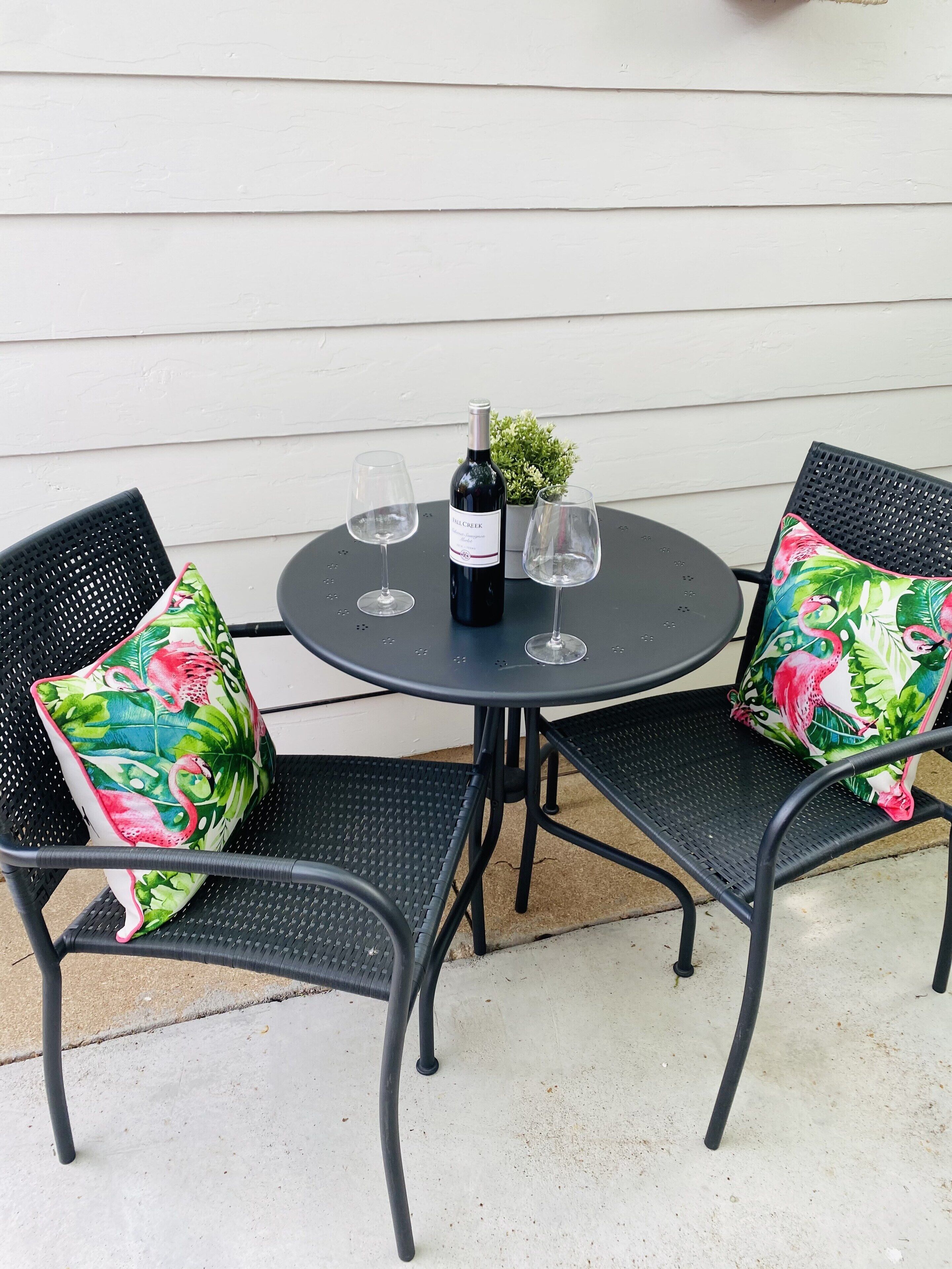 Een ronde tafel met twee stoelen, elk met een bloemenkussen. Op de tafel staat een fles wijn en twee glazen.
