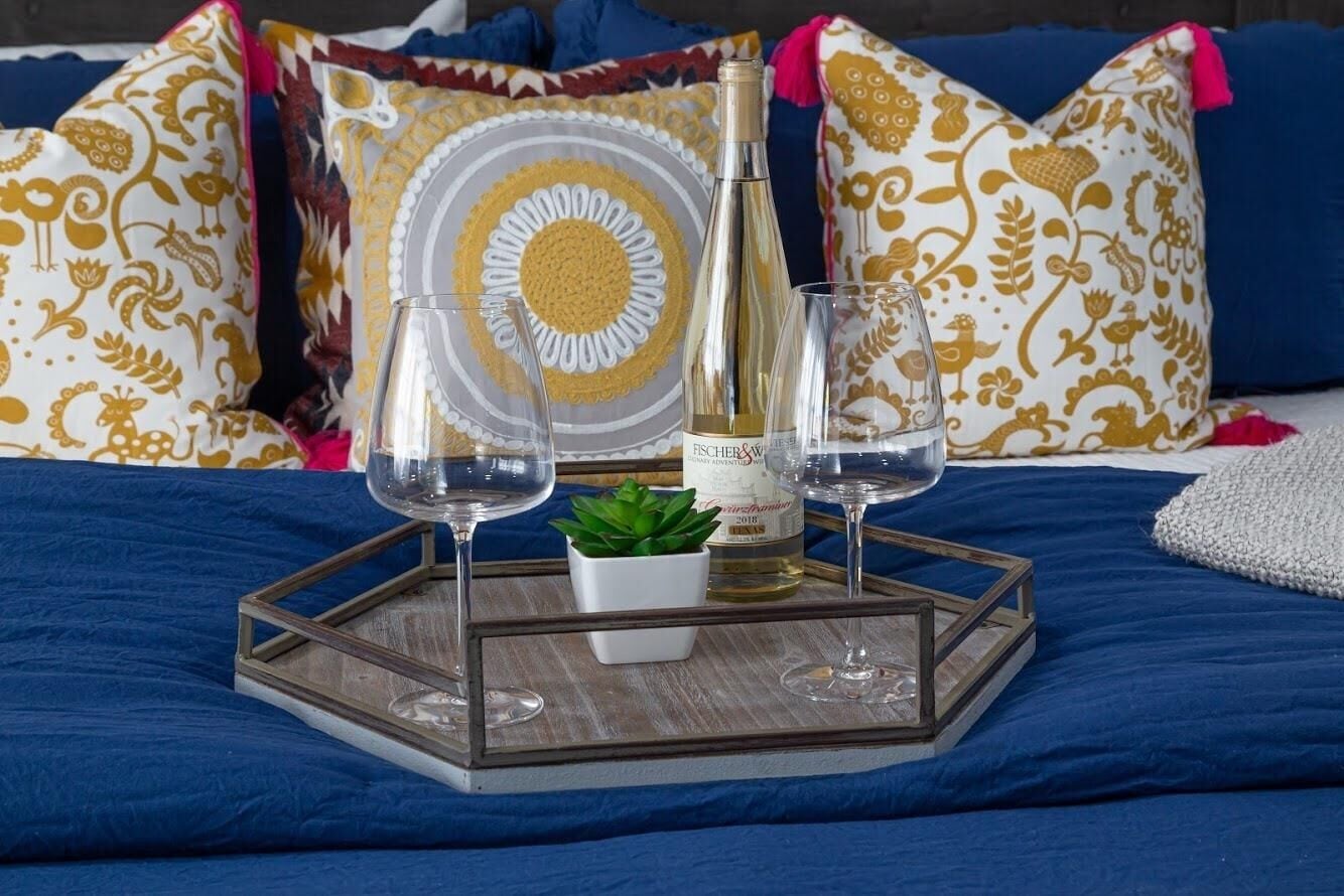 Una cama con una colcha azul, una bandeja decorativa con copas de vino, una botella y una pequeña planta.