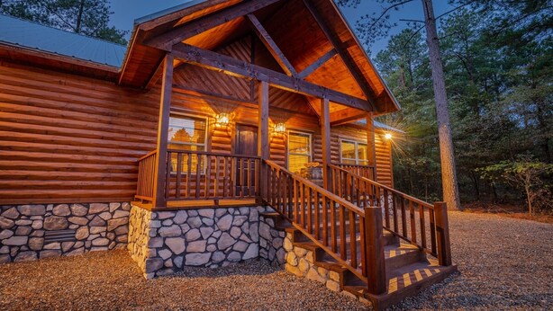 A log cabin with a stone foundation, a wooden porch with a staircase, and a view of the surrounding forest. - Romantic Music-Inspired Cabin with Hot Tub, Spa Bath, Outdoor TV & Fireplaces (Broken Bow)