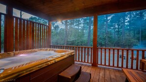 A hot tub on a wooden deck with a view of a forest. - Romantic Music-Inspired Cabin with Hot Tub, Spa Bath, Outdoor TV & Fireplaces (Broken Bow)