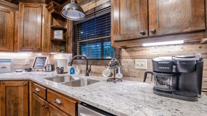 A kitchen with wooden cabinets, a granite countertop, a Keurig coffee maker, and a window with blinds. - Romantic Music-Inspired Cabin with Hot Tub, Spa Bath, Outdoor TV & Fireplaces (Broken Bow)