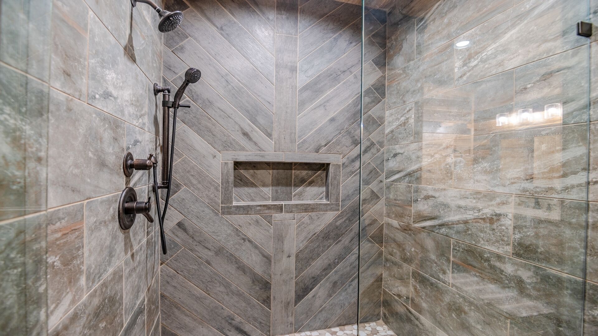 A shower area with a herringbone tile pattern, a handheld showerhead, and a wall-mounted showerhead.