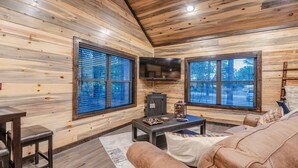 A cozy cabin interior with wooden walls, a fireplace, a sofa, and large windows. - Romantic Music-Inspired Cabin with Hot Tub, Spa Bath, Outdoor TV & Fireplaces (Broken Bow)