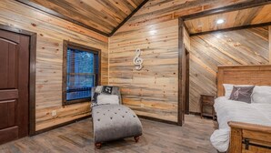 A bedroom with a wooden sloped ceiling, a bed, a small seating area, and a window with blinds. - Romantic Music-Inspired Cabin with Hot Tub, Spa Bath, Outdoor TV & Fireplaces (Broken Bow)