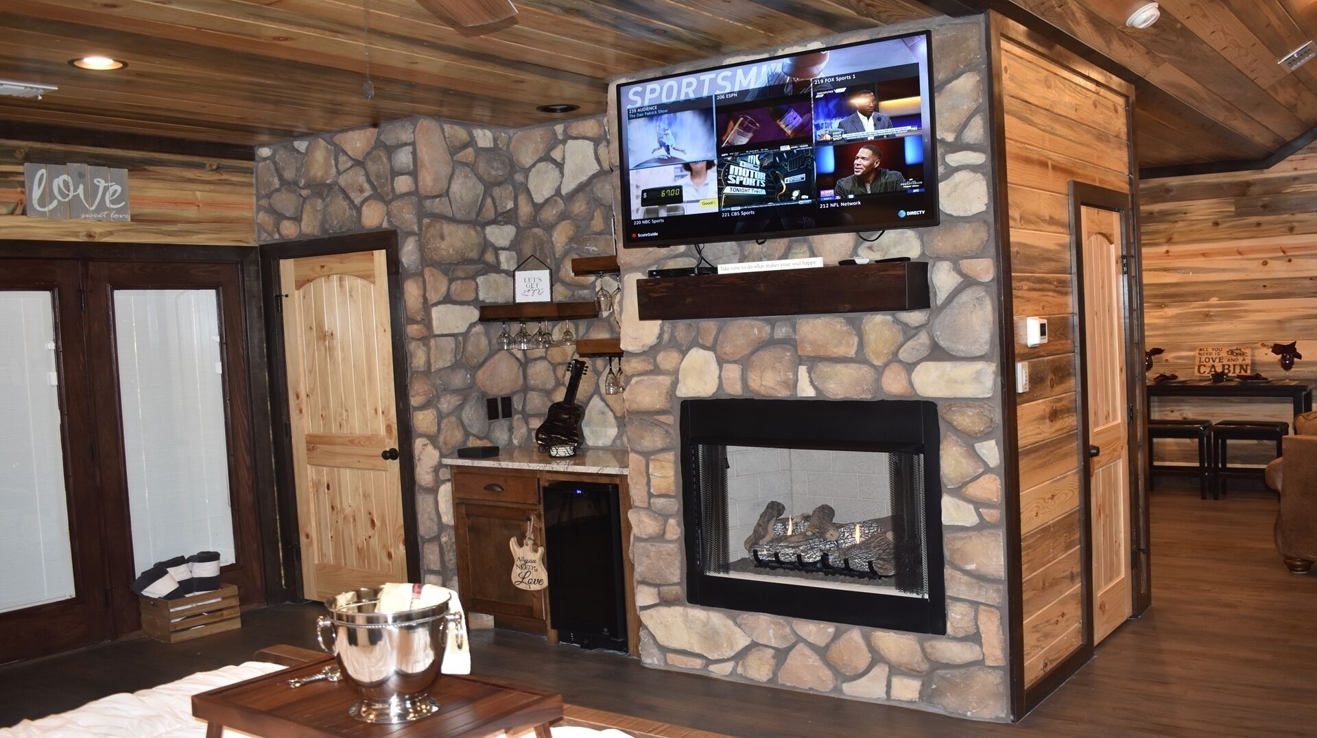 A cozy cabin interior with a stone wall, a fireplace, a television mounted on the wall, and a wooden door.