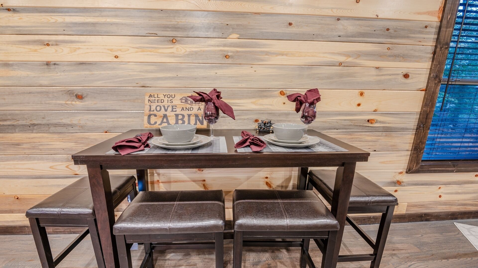 L’intérieur d’un chalet en bois, avec une table à manger dressée pour deux personnes, deux bols blancs, deux verres à vin et une pancarte sur laquelle on peut lire : « TOUT CE DONT VOUS AVEZ BESOIN, C’EST L’AMOUR ET UN CHALET ».