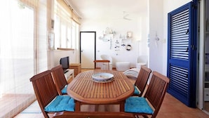 Zona pranzo con tavolo rotondo in legno e sedie, una ventola a soffitto e vista sul mare attraverso ampie finestre.