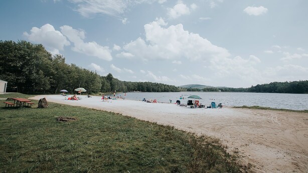 A lakeside beach with picnic tables and umbrellas, surrounded by trees and hills.
