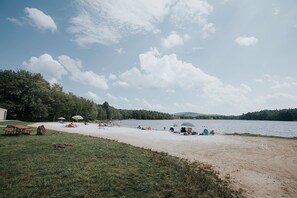 A lakeside beach with picnic tables and umbrellas, surrounded by trees and hills.