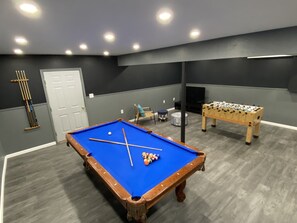 A pool table with balls, a foosball table, a television, and a seating area with a chair and ottoman.