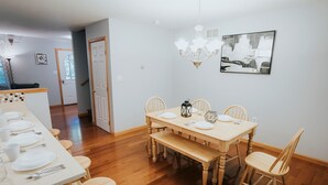 A dining area with a wooden table set for four, surrounded by wooden chairs. A chandelier hangs above the table, and a black lantern is placed on the table.