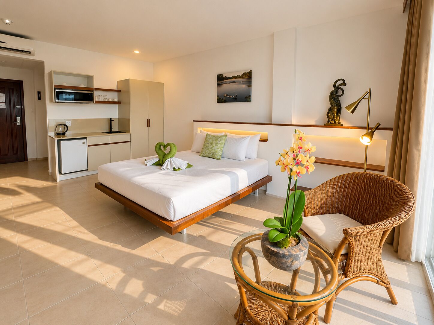 Quarto de hotel com uma cama, uma pequena cozinha, uma cadeira de vime, uma mesa de vidro e uma planta em vaso.
