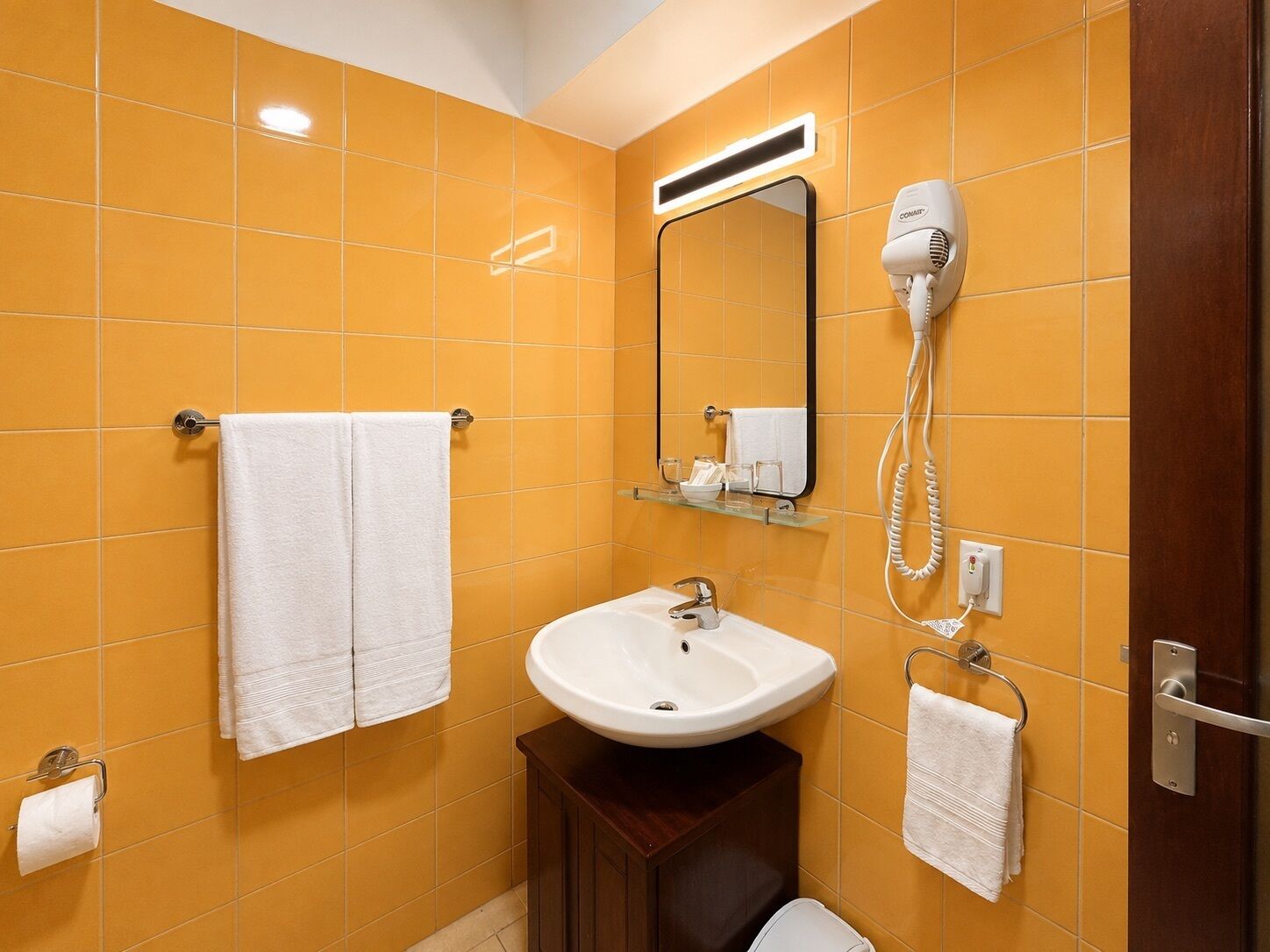 A bathroom with yellow tiles, a white sink, a mirror, a hairdryer, and a towel rack.