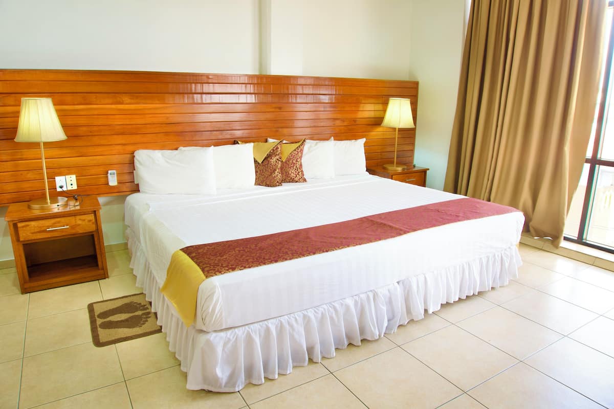 Habitación de hotel con una cama grande, cabecera de madera, dos mesitas de noche con lámparas y vistas al exterior a través de una ventana con cortinas.
