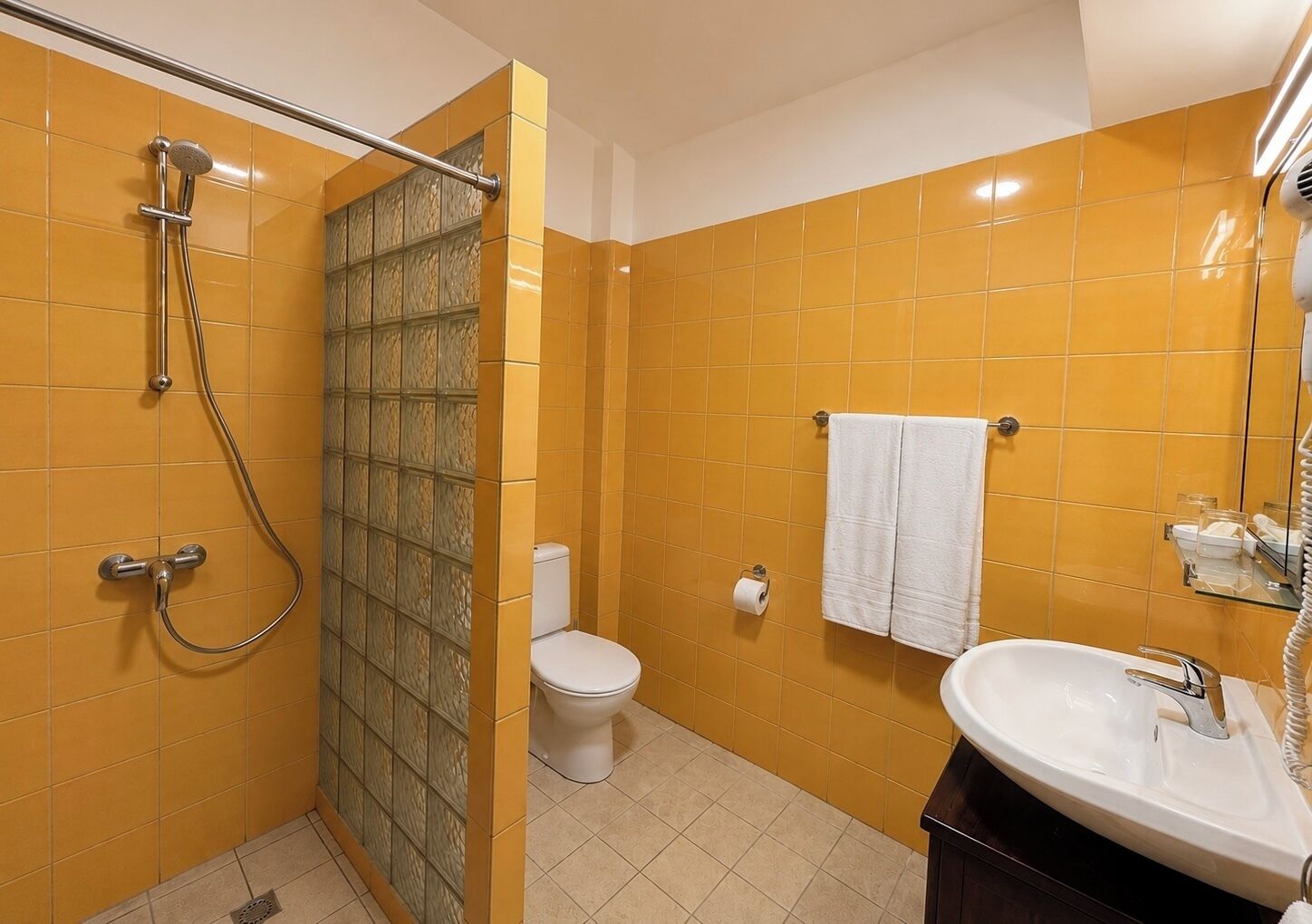 Un baño con baldosas amarillas, cabina de ducha de vidrio, inodoro, lavamanos y un perchero para toallas.