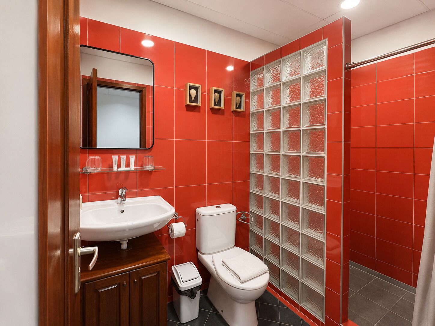 Un baño con pared de azulejos rojos, un lavabo blanco, un inodoro y una cabina de ducha con paredes de vidrio.