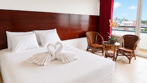 A hotel room with a large bed, a wooden headboard, a balcony with wicker chairs, and a view of buildings. - Spanhoek Boutique Hotel (Paramaribo)