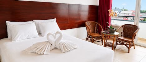 A hotel room with a large bed, a wooden headboard, a balcony with wicker chairs, and a view of buildings.