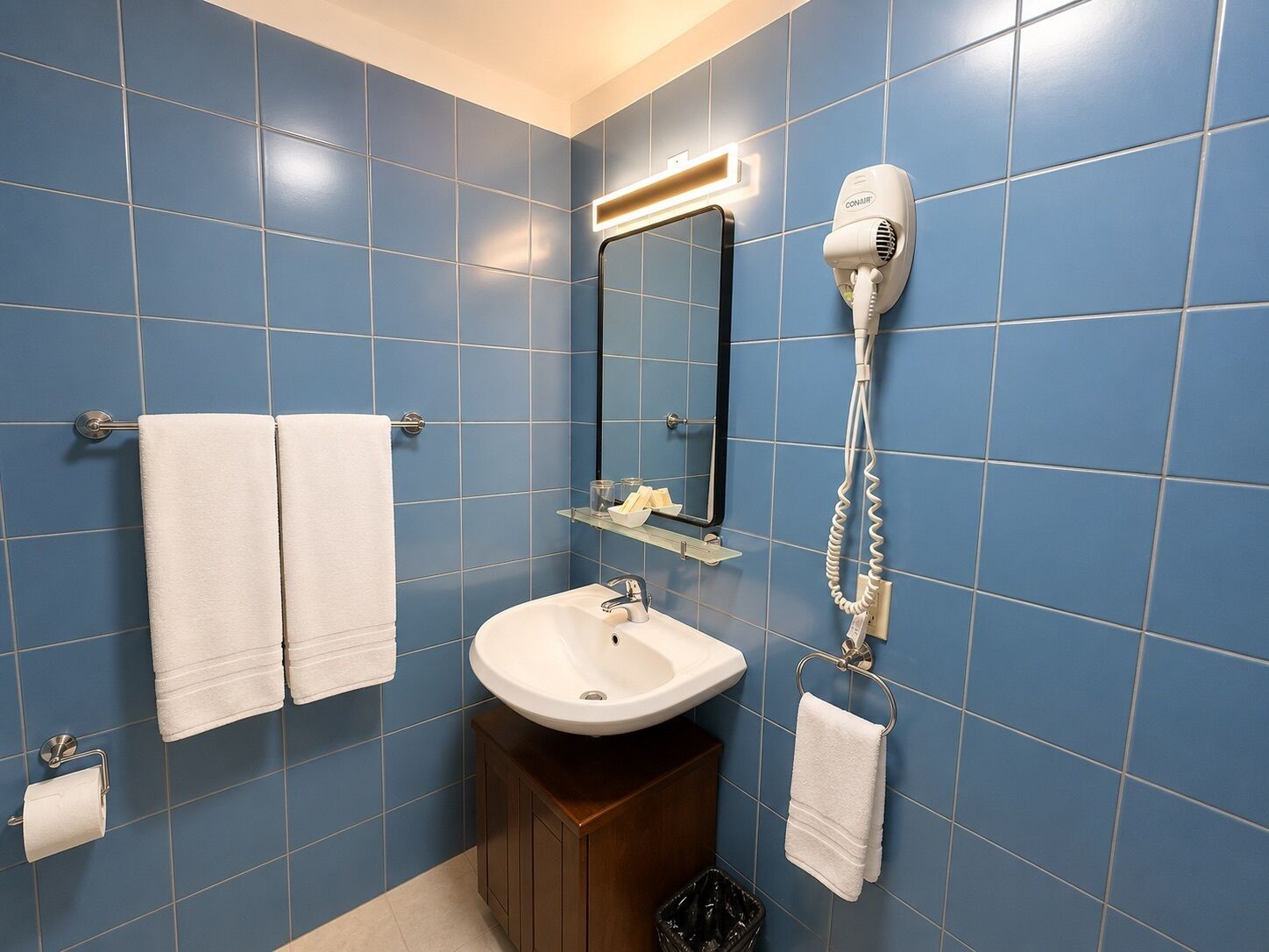 Un baño con azulejos azules, un lavamanos blanco, un secador de pelo y dos toallas.
