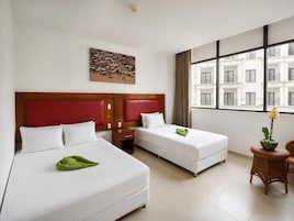 Habitación de hotel con dos camas, un cuadro en la pared, un ventanal con vista a edificios y una mesita con una planta.