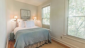A bedroom with a bed, two bedside lamps, a window with blinds, a rug, and a wall-mounted artwork. - Historic Cottage with modern amenities (louisville)