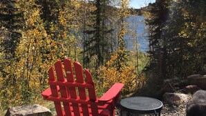 A red Adirondack chair and a small round table on a gravel surface, surrounded by autumn foliage and a lake view. - Lakeside Adventurer's Cabin (Durango)