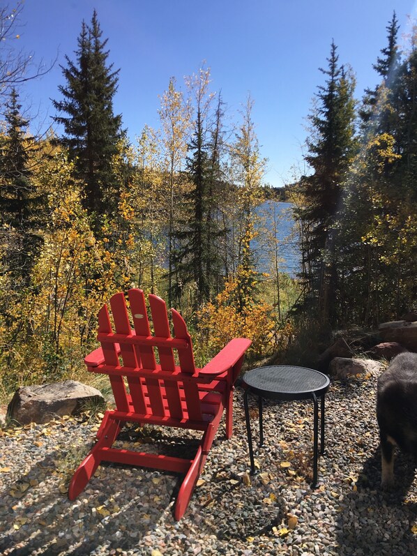 A red Adirondack chair and a small round table on a gravel surface, surrounded by autumn foliage and a lake view. - Lakeside Adventurer's Cabin (Durango)