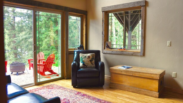 A room with a large sliding glass door leading to an outdoor area with red Adirondack chairs and a fire pit. There is a black leather chair with a decorative pillow, a wooden chest, and a framed picture on the wall. - Lakeside Adventurer's Cabin (Durango)