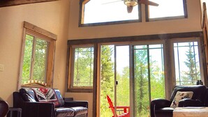 A spacious living room with a high ceiling, wooden beams, a ceiling fan, and large windows offering a view of the outdoors. - Lakeside Adventurer's Cabin (Durango)