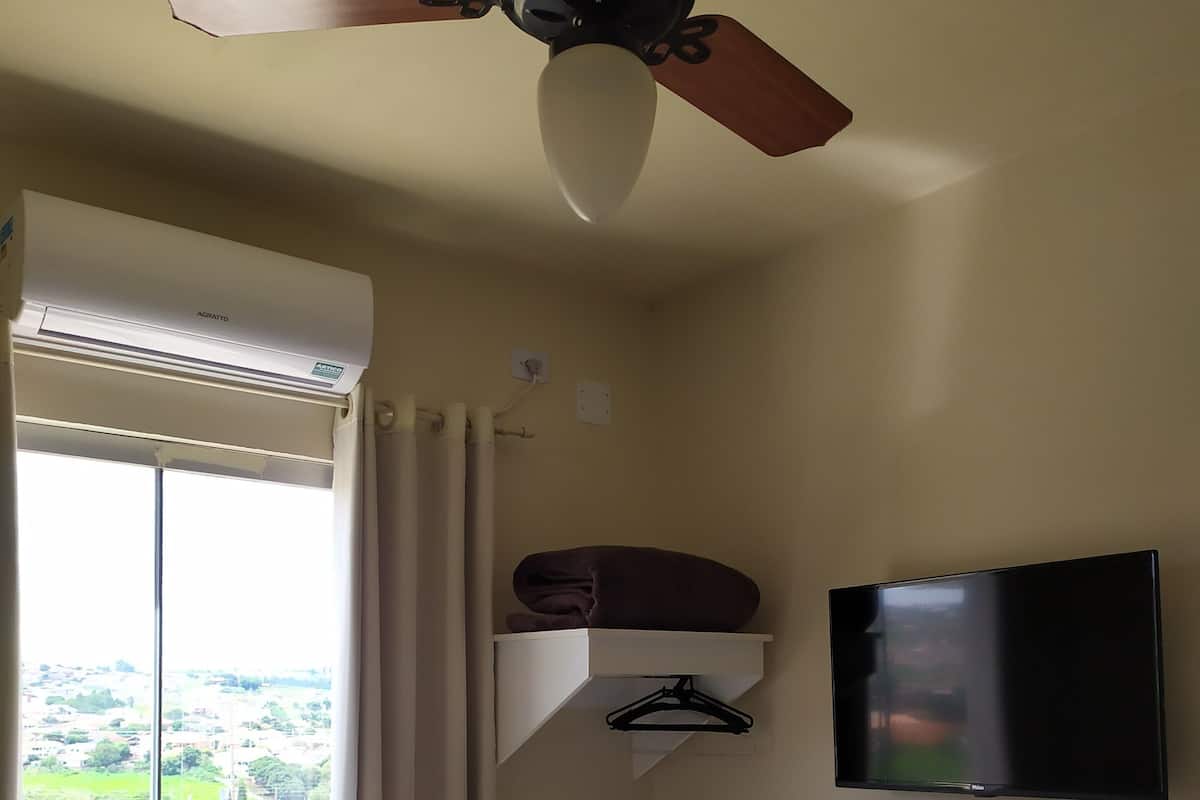A hotel room with a bed, a ceiling fan, a TV mounted on the wall, a window with curtains, and a view of the outside.