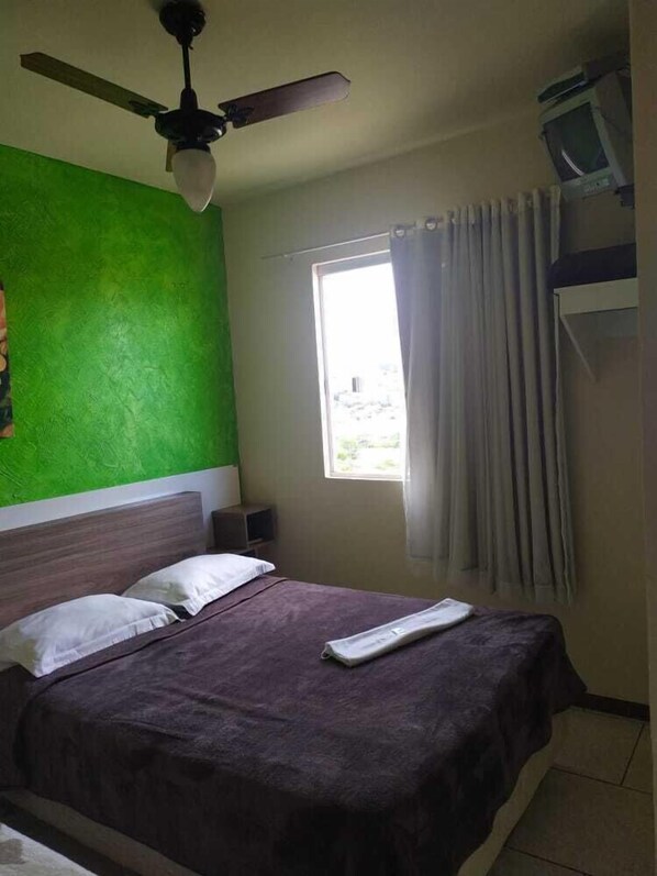 Ein Schlafzimmer mit einem Bett, einem Deckenventilator, einem Fenster mit Vorhängen und einem Fernseher auf einem Schrank.