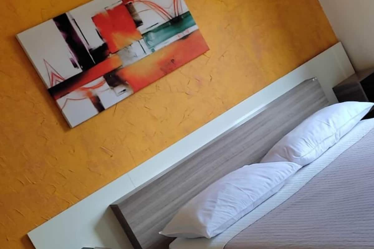 A hotel room with a bed, a bedside table, a nightstand, a wall-mounted air conditioner, and a large abstract painting on the wall.