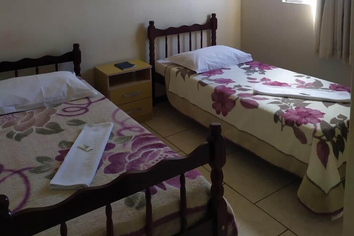 A room with two single beds, a ceiling fan, a window with curtains, and a small bedside table with a lamp.