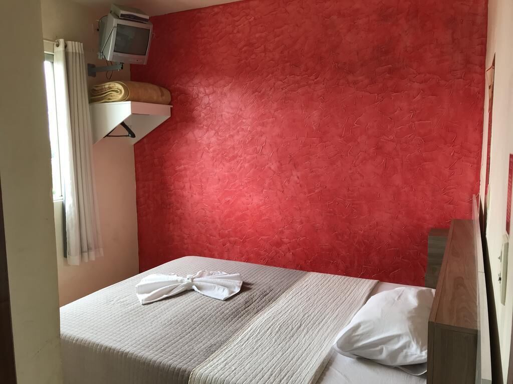 A small, neatly made bed with a white bedspread and a white pillow, a red textured wall, a small shelf with a television and some items, and a window with curtains.
