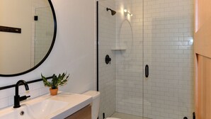 Ein Badezimmer mit Waschbecken in Weiß, einem runden Spiegel, einer Topfpflanze, einer Duschkabine mit Glastür und einer weißen Toilette.