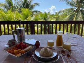 Ein Tisch ist reichhaltig mit Frühstücksartikeln gedeckt, darunter Obst, Reis und Getränke, mit Blick auf Palmen.