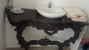 Um banheiro com pia branca, toucador escuro ricamente decorado, espelho e um vaso com flores.