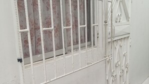 Paredes externas brancas com uma janela com barras brancas e um portão de metal decorativo. Vasos com plantas de folhas variegadas ao longo da base da parede.