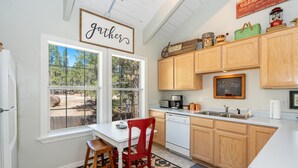 A kitchen with wooden cabinets, a white countertop, a window with a view of trees, and a 'Welcome' sign on the wall. - Magdalena Cottage on 1/2 acre (Flagstaff)