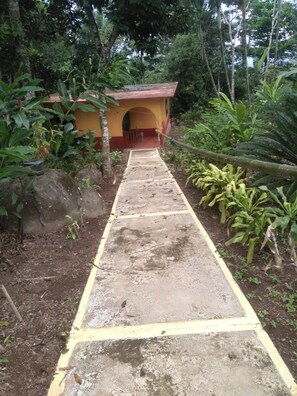 Un camino de concreto lleva a una pequeña construcción amarilla con techo rojo, rodeada de follaje.